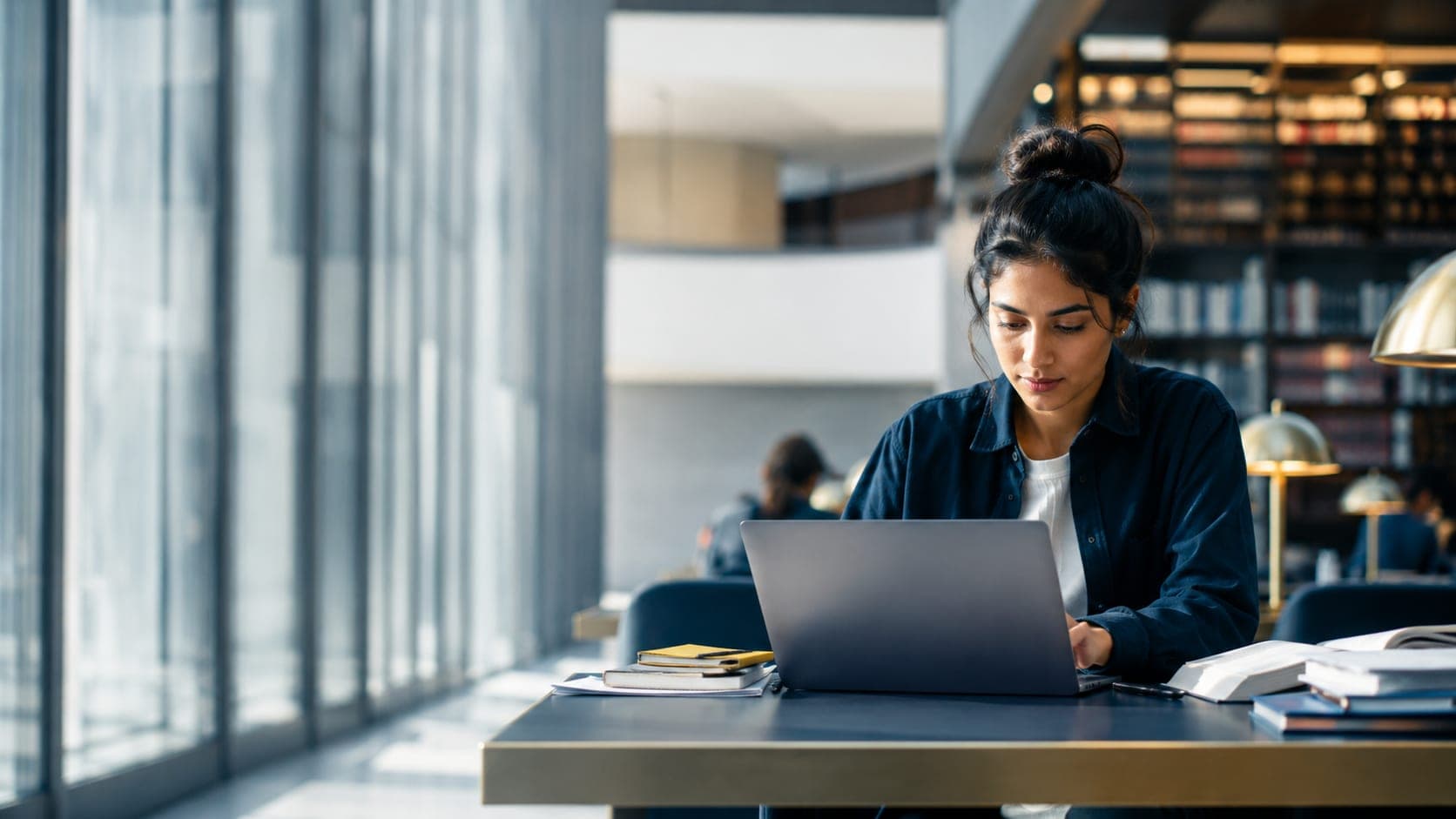 A student studying on a laptop in a bright university library.